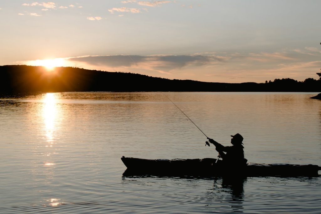pexels photo 5688674 Side view of anonymous male silhouette in boat catching fish with rod on rippled river under cloudy sky with shiny sun at dusk